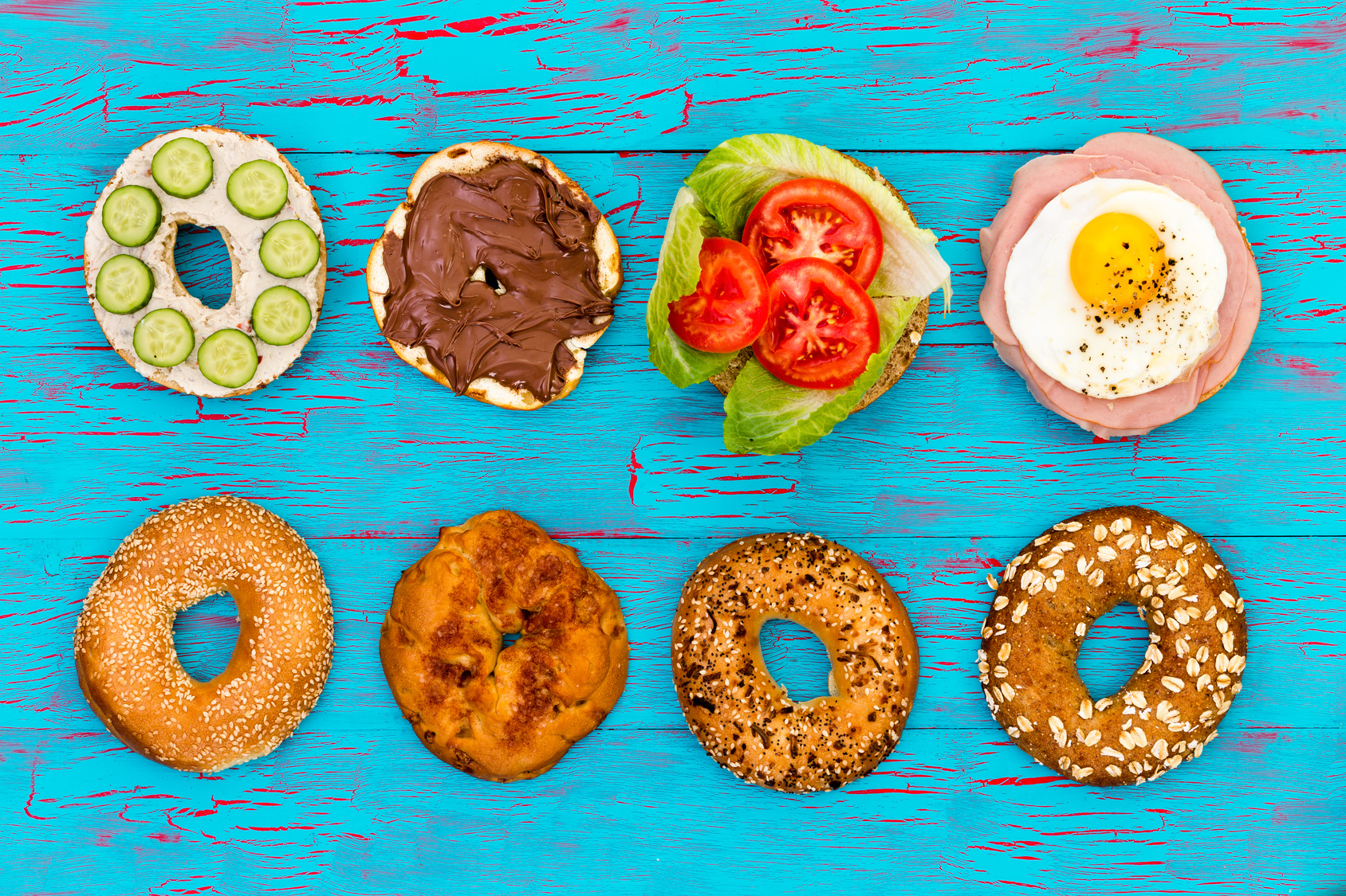 Four different flavored fresh sliced bagels with assorted filling of cucumbers, chocolate , salad, fried egg and ham, neatly arranged in a row on an exotic blue picnic table viewed from above