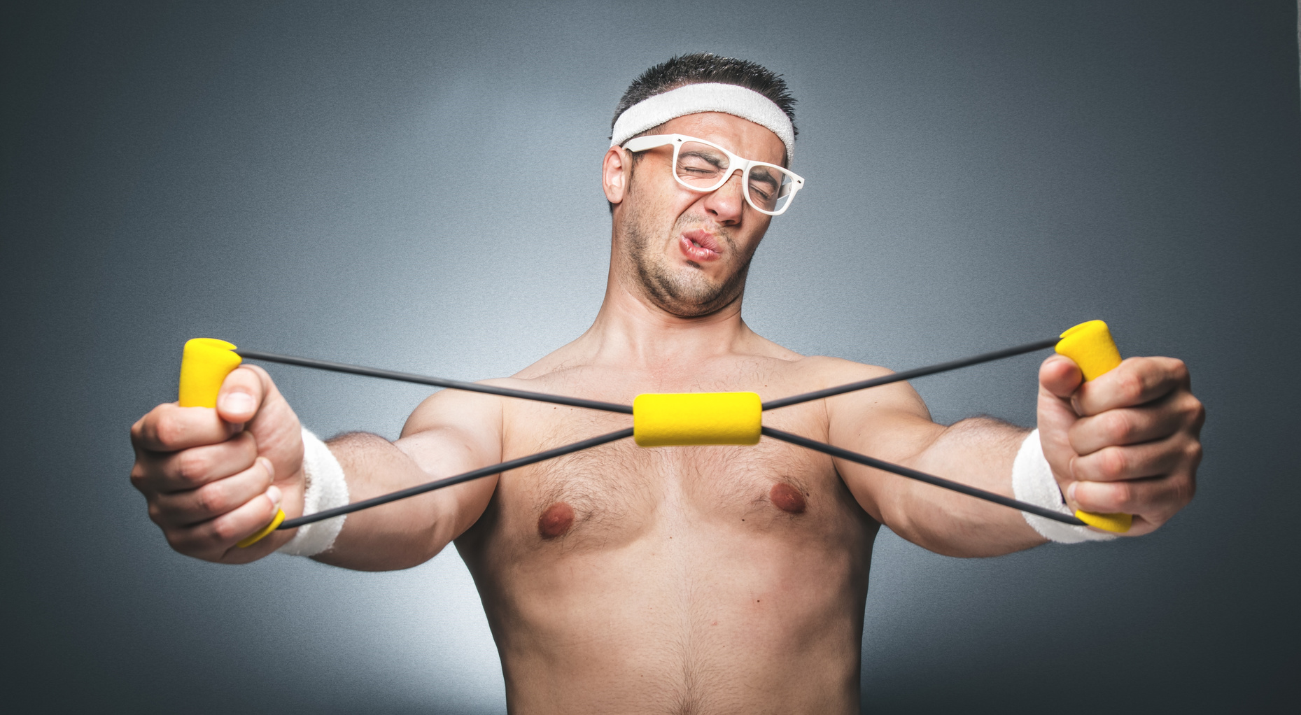 Silly man exercising over dark gray background. Nerd guy with eyeglasses holding expander. Close up of funny young man doing aerobics. Studio shot