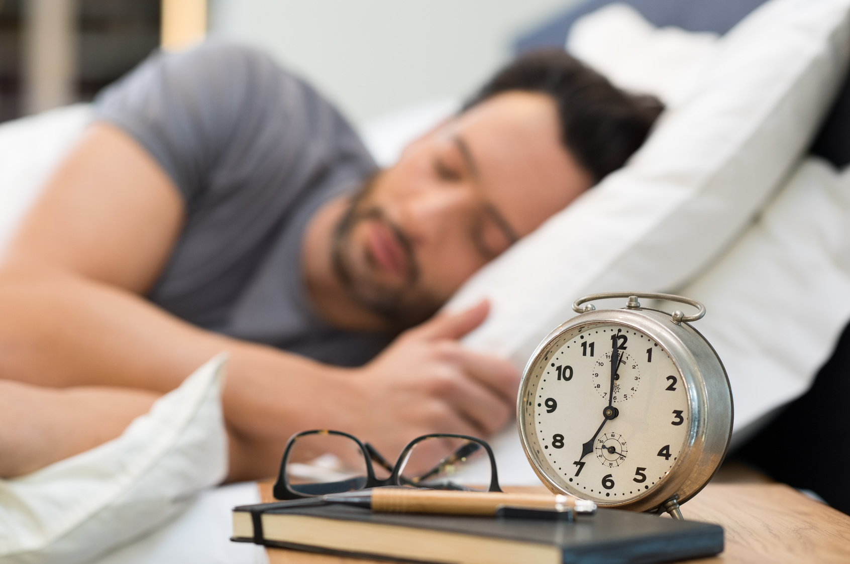 Young man sleeping in his bedroom. Man sleeping with an alarm clock in foreground. A calm man in his bed before waking up in his room. Close up of alarm clock on bedside table.