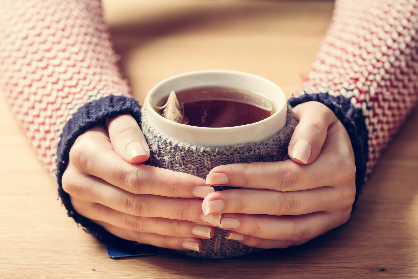 Hot mug of tea warming woman's hands in retro woollen jumper. Wooden table