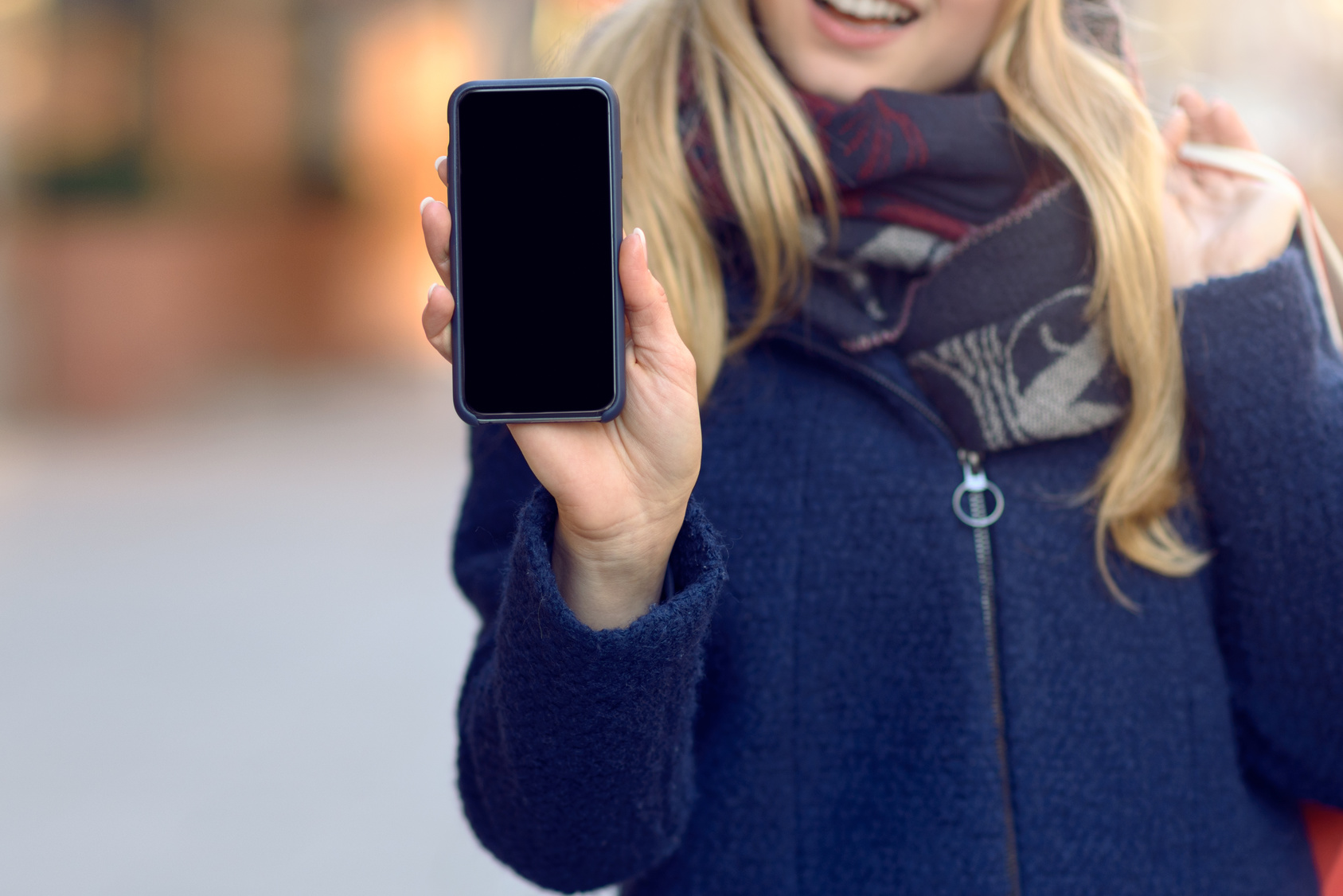 Pretty young woman holding up her mobile phone with a blank display as she stands on an urban street in winter warmly dressed in a knitted woollen scarf, hat and jacket