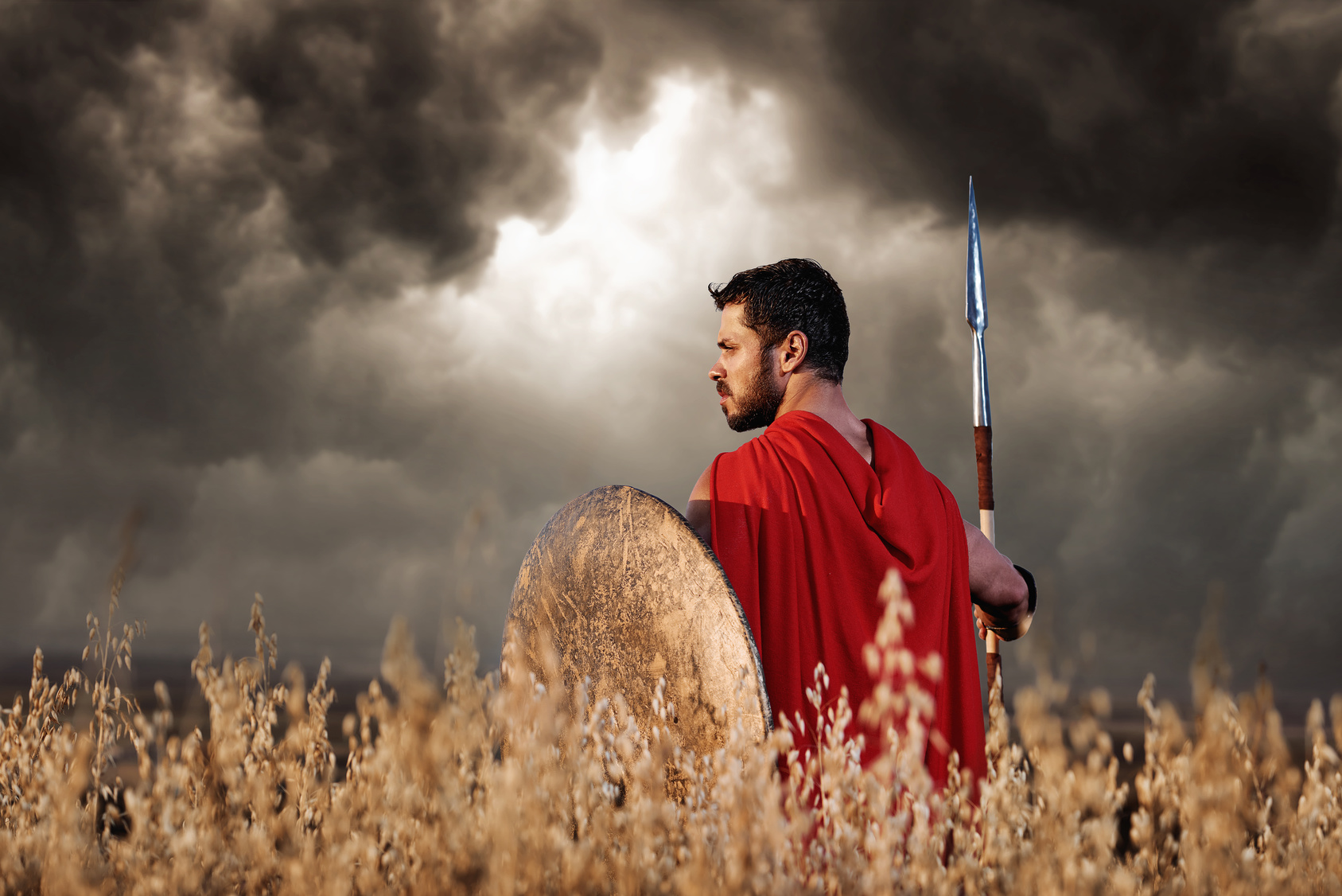 Back view of warrior wearing in red cloak like spartan or antique roman solider turned back and looking away. Brunet with beard holding sword and shield going in attack. Bad weather with dark clouds.