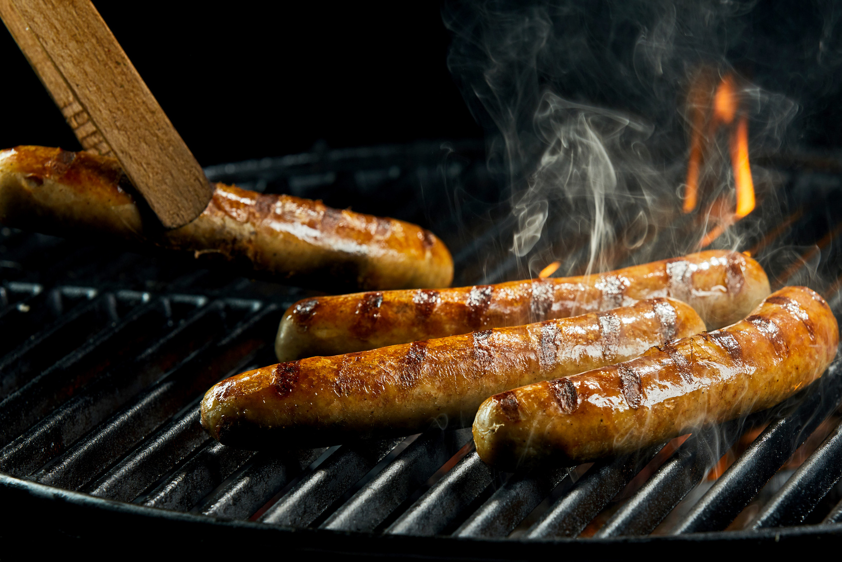 Succulent sausages grilling on a BBQ fire with flames and smoke or steam and wooden tongs turning one over