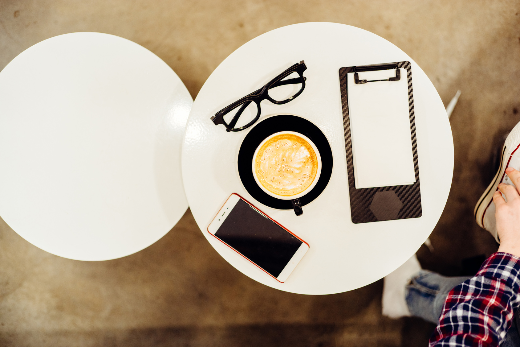 Top view of glasses, cup of cappuccino, clipboard with blank paper and smartphone  in modern cafe interior.